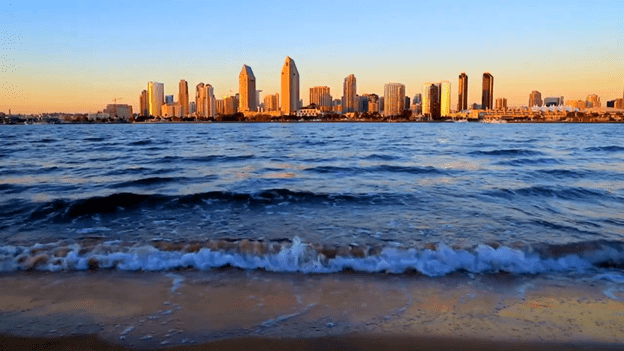 The surf rolling in on a beach with the San Diego skyline at sunset behind it.